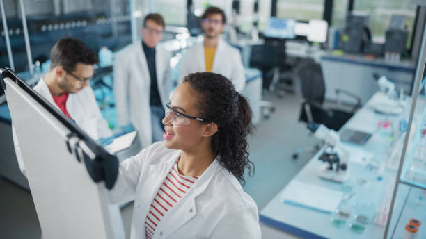 istock Medical Science Laboratory: Beautiful Black Female Scientist Writes Detailed Project Data Analysis on the Board, Her Diverse Team of Colleague Listens. Young Scientists Solving Problems. 1346677602