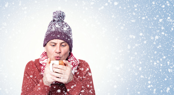 Portrait of a man in a red sweater, scarf and hat, holding a hot drink in a mug and blowing on it. On a blue background with snow