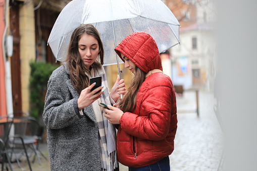 Two girls using smart phones during a bad weather condition in the city
