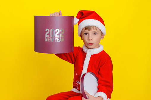 Smiling six-year-old boy dressed as Santa Claus holding a round red gift box with the inscription New Year 2022, close-up studio portrait