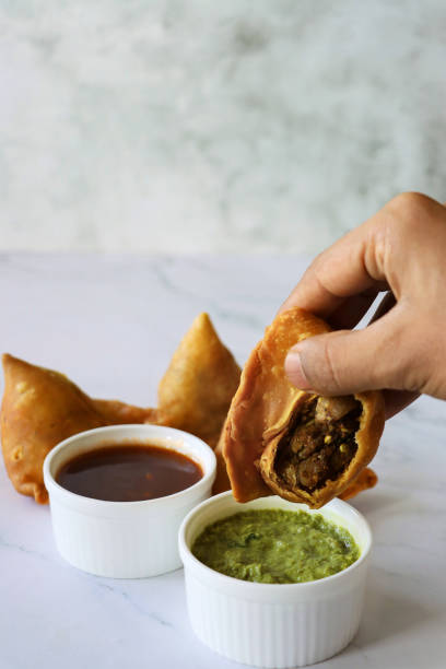 istock Image of unrecognisable person dipping a fried samosa with missing bite, stuffed with spiced potato, peas and meat into ramekin of mint coriander dip and mango chutney, on a marble effect background, elevated view, focus on foreground 1346687792