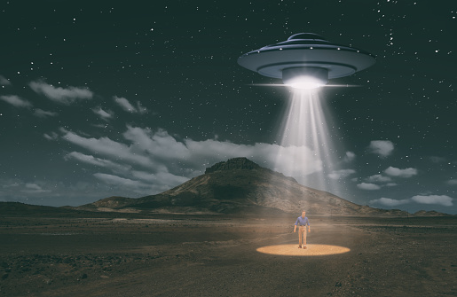 An old-style UFO hovers over a desert landscape at night, probing the ground with bright lights. Standing in the light beam is a humanoid figure, though is he human or alien..? Miniature photography.