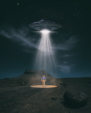 An old-style UFO hovers over a desert landscape at night, probing the ground with bright lights. Standing in the light beam is a humanoid figure, though is he human or alien..? Miniature photography.