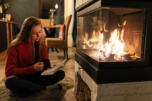 Teenage girl enjoying looking at the fire burning in the fireplace. The girl is reading a book and drinking a cup of hot chocolate.
Shot with Canon R5