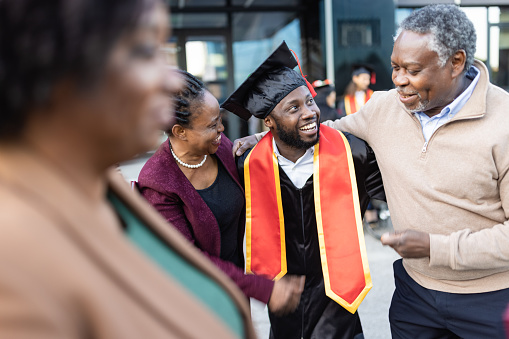 Excited African American college graduate smiles and talks with his mom and dad after the graduation ceremony.