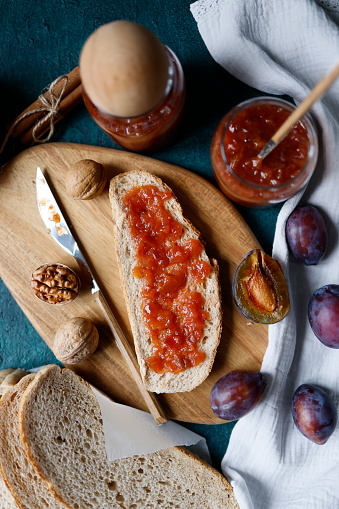 Istanbul, Turkey-October 10, 2021: Damson plum marmalade spread on slices of brown wheat bread on wooden cutting board on dark turquoise green rough concrete floor. There is damson plums, slices of bread, two jars of marmalade, walnuts and a gray linen napkin.