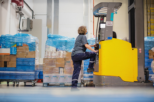 Female warehouse worker in protective work-wear getting onto pallet truck to start loading stored boxes on stack