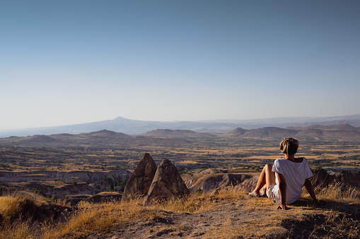 rear view of blonde Caucasian young woman with white shirt and shorts sitting on a hill and admiring the beautiful valley landscape of Cappadocia Turkey