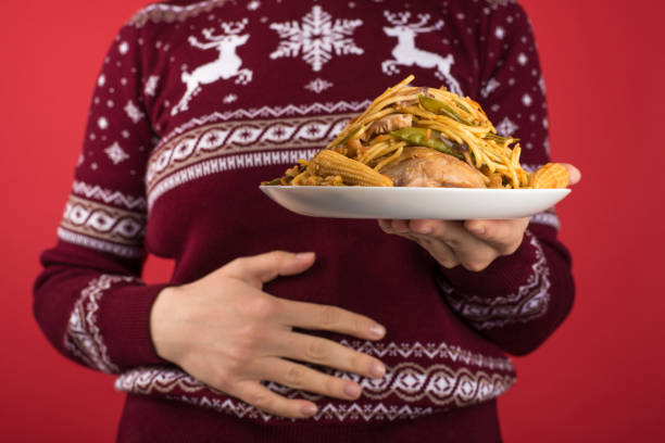 Cropped closeup photo of young woman in red and white christmas sweater holding large plate of fatty food and touching her hurting stomach on isolated red background Cropped closeup photo of young woman in red and white christmas sweater holding large plate of fatty food and touching her hurting stomach on isolated red background big plate of food stock pictures, royalty-free photos & images