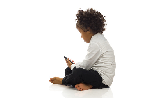 Little black boy in elegant clothes is sitting on a floor, holding telephone and looking at it. Side view. Full length studio shot isolated on white.