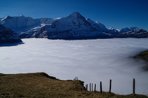 A spectacular series of 5 images recording an especially early incidence of Temperature Inversion in the Grindelwald Valley of the Bernese Oberland in Switzerland, October 2021. Such meteorological events are more common in the depths of winter than early autumn, making these images unique. The extreme, acute angle of the light at this time of year makes these images stand out from the crowd. They are all taken from Grindelwald First (2166m/7009ft) which is reached by one of the world's longest Gondola Cable Cars in the world. he cloud top on this October day in 2021 was at around 6000ft with the base at ground level, closer sea level but not at zero. Temperature inversion results in warm air sitting on top of cold air and keeping the cloud hanging in valleys when in mountainous areas. In these images only the very tallest peaks of the Bernese Oberland can be seen to emerge. The two Alpine massifs clearly visible are (1) in the Haslital Region, specifically Mount Titlis and its surrounding peaks (Images 1 & 4) and in the Jungfrau Region, specifically the Eiger, Monch and Jungfrau (Images 2, 3 & 5). Specifically each image is as follows:\nImage 2: The Eiger, Monch & Jungfrau and surrounding peaks with the North Face of the Eiger completely in shadow but emerging from the sea of cloud. The fence pots marl the boundary of the land into the abyss.
