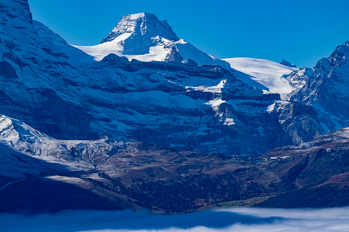A spectacular series of 5 images recording an especially early incidence of Temperature Inversion in the Grindelwald Valley of the Bernese Oberland in Switzerland, October 2021. Such meteorological events are more common in the depths of winter than early autumn, making these images unique. The extreme, acute angle of the light at this time of year makes these images stand out from the crowd. They are all taken from Grindelwald First (2166m/7009ft) which is reached by one of the world's longest Gondola Cable Cars in the world. he cloud top on this October day in 2021 was at around 6000ft with the base at ground level, closer sea level but not at zero. Temperature inversion results in warm air sitting on top of cold air and keeping the cloud hanging in valleys when in mountainous areas. In these images only the very tallest peaks of the Bernese Oberland can be seen to emerge. The two Alpine massifs clearly visible are (1) in the Haslital Region, specifically Mount Titlis and its surrounding peaks (Images 1 & 4) and in the Jungfrau Region, specifically the Eiger, Monch and Jungfrau (Images 2, 3 & 5). Specifically each image is as follows:\nImage 3: The Jungfrau & Tschingelhorn above Kleine Scheidegg, clearly visible below the snow line.