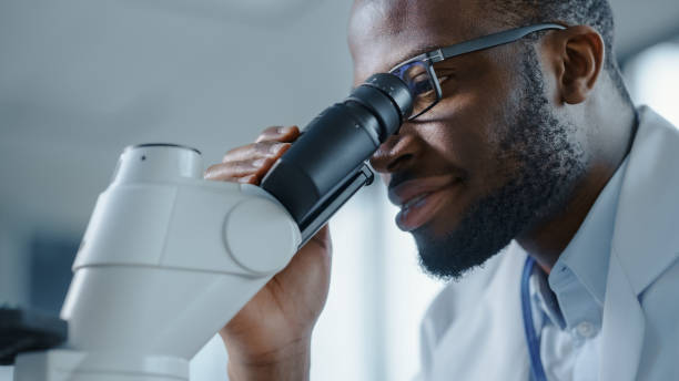 foto de primer plano macro de un apuesto científico negro con gafas y mirando hacia el microscopio. microbiólogo trabajando en muestras de moléculas en laboratorio moderno con equipos tecnológicos. - medical research fotos fotografías e imágenes de stock