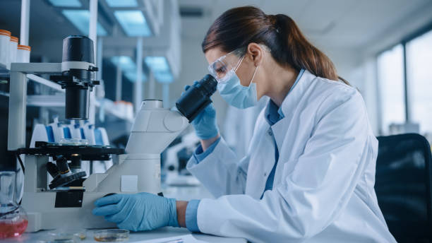 istock Female Scientist in Face Mask and Glasses Looking a Petri Dish with Genetically Modified Sample Chemicals Under a Microscope. Microbiologist Working in Modern Laboratory with Technological Equipment. 1346705954