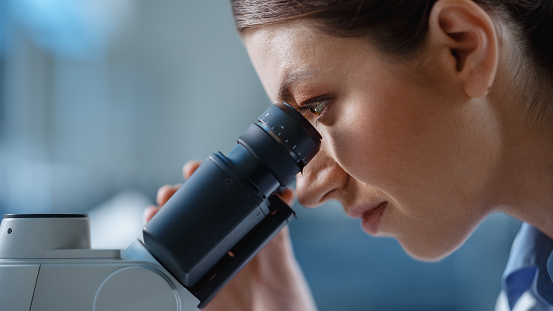 Macro Close-Up Shot of a Beautiful Female Scientist Looking into the Microscope. Woman Microbiologist Working on Molecule Samples in Modern Laboratory with Technological Equipment.