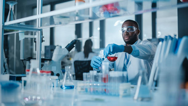 scienziato maschio afroamericano che indossa occhiali protettivi che mescolano sostanze chimiche in una provetta in un laboratorio. bel microbiologo nero che lavora in un laboratorio moderno con attrezzature tecnologiche. - laboratory test tube student scientist foto e immagini stock