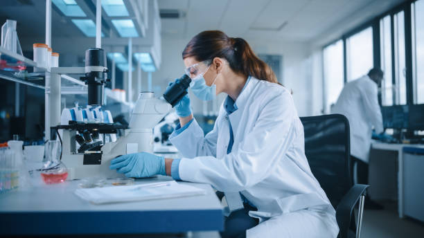 istock Female Scientist in Face Mask and Glasses Looking a Petri Dish with Genetically Modified Sample Chemicals Under a Microscope. Microbiologist Working in Modern Laboratory with Technological Equipment. 1346706046