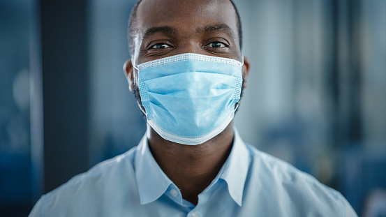 Close Up Portrait of a Black African American Handsome Male in White Shirt Wearing a Protective Face Mask. Successful Man Calmly Looking at Camera. Stylish Businessman or a Doctor in Hospital.