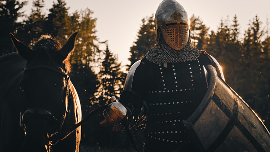 Medieval Knight Warrior Leading His Horse into the Battle to Fight. Portrait of Hero in Plated Armor, Shield, Helmet, Sword and Thoroughbred Stallion. Cinematic Light, War, Smoke, Invasion, Conquest