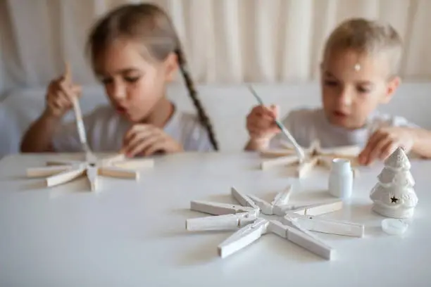 Siblings, boy and girl, paint wooden snowflakes from clothespins with white paint at home, hands in paint, recyclable craft ornament, New Year DIY decoration, selective focus on snowflake