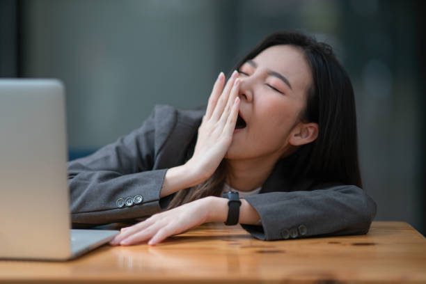 junge geschäftsfrau gähnt an einem modernen schreibtisch vor einem laptop und bedeckt aus höflichkeit ihren mund - yawning stock-fotos und bilder