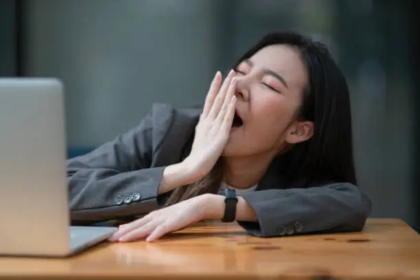 Young businesswoman yawning at a modern office desk in front of a laptop, covering her mouth out of courtesy