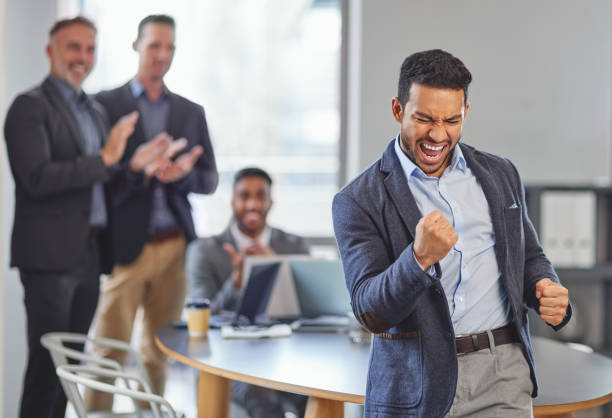 shot of a young businessman celebrating a win in front of his colleagues having a meeting in the background - excitement business person ecstatic passion imagens e fotografias de stock