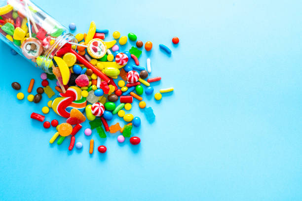 Glass jar spilling candies on blue background Large group of all sort of multicolored candies spilling out from a glass jar shot from above on blue background. High resolution 42Mp studio digital capture taken with SONY A7rII and Zeiss Batis 40mm F2.0 CF lens gummi bears photos stock pictures, royalty-free photos & images