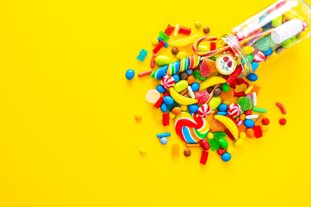 Glass jar spilling candies on yellow background Large group of all sort of multicolored candies spilling out from a glass jar shot from above on yellow background. High resolution 42Mp studio digital capture taken with SONY A7rII and Zeiss Batis 40mm F2.0 CF lens candy jellybean variation color image stock pictures, royalty-free photos & images