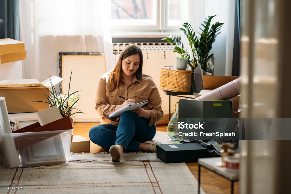 Moving Out: Beautiful Smiling Overweight Woman Sitting on the Floor Surrounded by Packed Boxes and Making a To-do List A happy plus size woman moving out, sitting on the floor and making a list of items packed Relocation Stock Photo