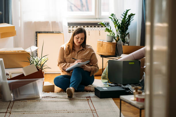 Moving Out: Beautiful Smiling Overweight Woman Sitting on the Floor Surrounded by Packed Boxes and Making a To-do List stock photo