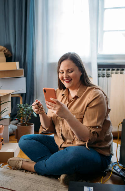 istock Moving in: Smiling Plus Size Woman Sitting on the Floor Browsing the Internet on her Smartphone and Taking a Break from Unpacking 1346727110