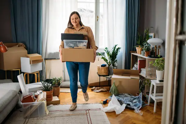 Photo of Ready to Move out: Happy Caucasian Plus Size Woman Standing in the Middle of a Messy Room while Holding Boxes with Personal Belongings