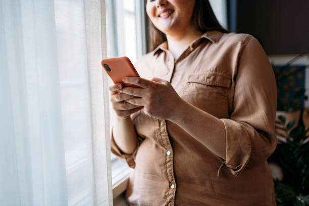 istock Unrecognizable Smiling Plus Size Woman Standing by the Window in her New Apartment and Using her Smartphone 1346727162