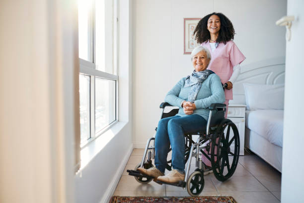 Full length portrait of an attractive senior woman sitting in a wheelchair with a female nurse crouching next to her in the retirement home stock photo