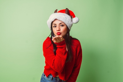 young latinx woman with santa claus hat, sweater and red lips blowing a kiss looking at camera.