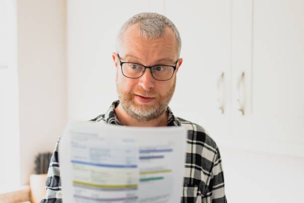 Worried man checking bills at home Portrait of a mid adult man checking his energy bills at home. He has a worried expression and touches his face with his hand while looking at the bills. Focus on the man while the interior architecture of the house is defocused. energy bill photos stock pictures, royalty-free photos & images