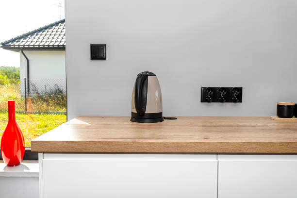 istock An electric kettle standing on the countertop on the kitchen cabinets visible a large window in the dining room and red vase. 1346741482