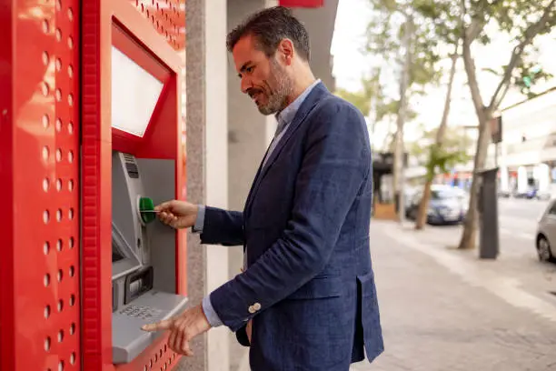 Side view of a well dressed successful middle aged businessman of Spanish ethnicity, standing next to the red colored ATM and using his credit card to withdraw his funds from his bank account