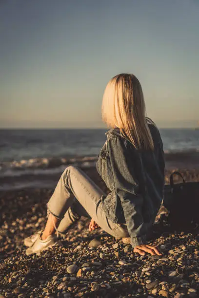 Photo of Young blonde woman is sitting on a pebble beach by the sea and looking at the sunset, a photo of her back. Concept of travel, enjoyment, loneliness. Vertical photo