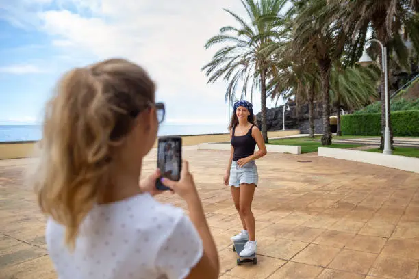 Two teenagers outdoors filming on the mobile phone a skate video to publish on social media accounts