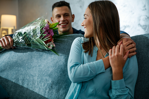 Candid shot of romantic young man standing behind the sofa, where his girlfriend is sitting, and surprising her with a beautiful bouquet of flowers for their anniversary.