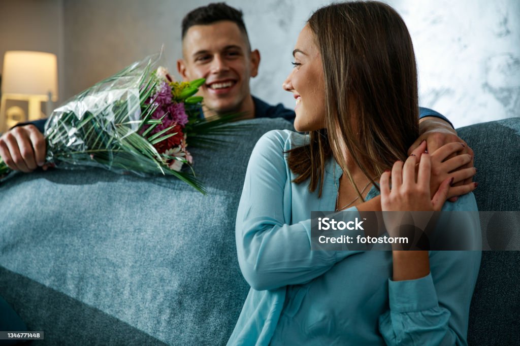 Romantic young man surprising his girlfriend with a bouquet of beautiful flowers Candid shot of romantic young man standing behind the sofa, where his girlfriend is sitting, and surprising her with a beautiful bouquet of flowers for their anniversary. Flower Stock Photo