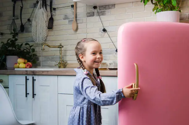 Photo of A five-year-old girl in a pink coat opens an old vintage pink refrigerator in a retro-styled kitchen