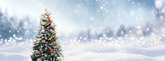Beautiful festive Christmas snowy background with holiday lights. Christmas tree decorated with red balls and knitted toys in forest in snowdrifts in snowfall on nature outdoors, panorama, copy space.