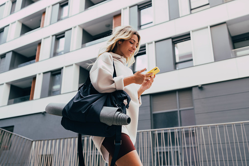 Cheerful smiling woman in sportswear typing text message on her smartphone while walking in the city