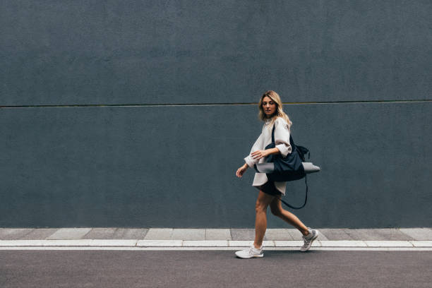 Sporty Woman in a White Sweatshirt Walking in City Young woman in sportswear with a sports bag and exercise mat walking down the street someone walking sneakers stock pictures, royalty-free photos & images