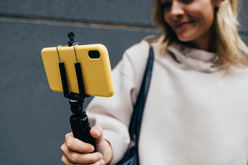 Close up photo of a female hand holding tripod stand with a yellow smartphone while standing over a gray wall