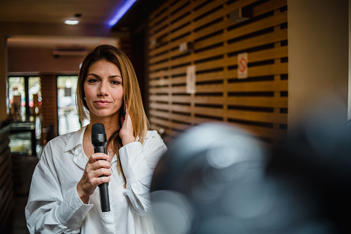 Portrait of a professional journalist at work. A young woman is standing in a cafe with a microphone in her hand and is ready to join the live TV program.