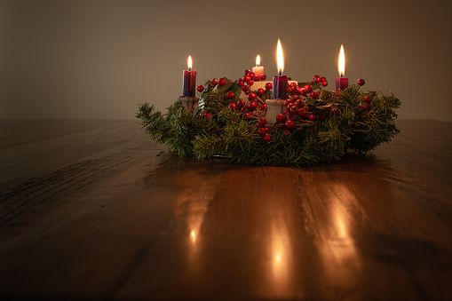 Advent wreath with evergreen boughs and red berries with four candles lit sitting on a dark wood table with candlelight reflecting off table shot from tabletop with copy space